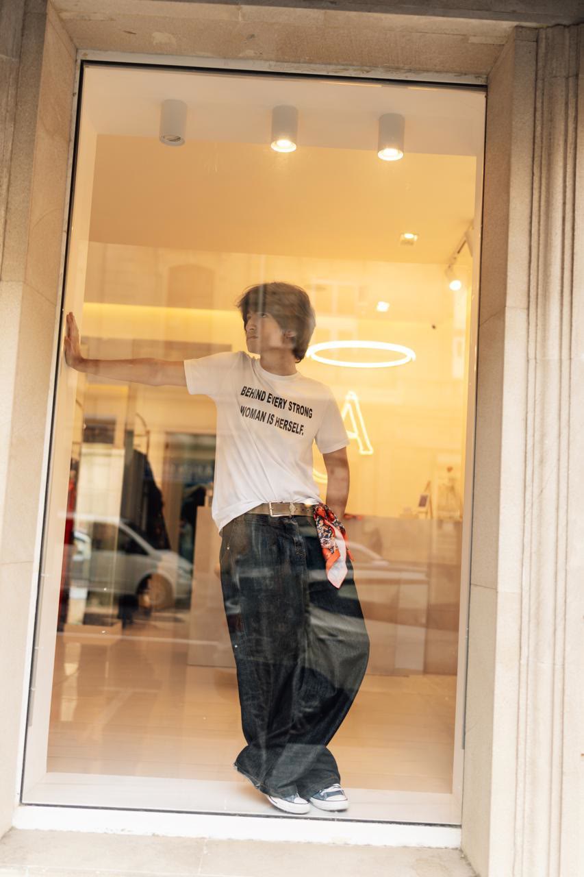 Jamal leans against the inside of a lit shop window in a white T-shirt that reads “Behind every strong woman is herself.”