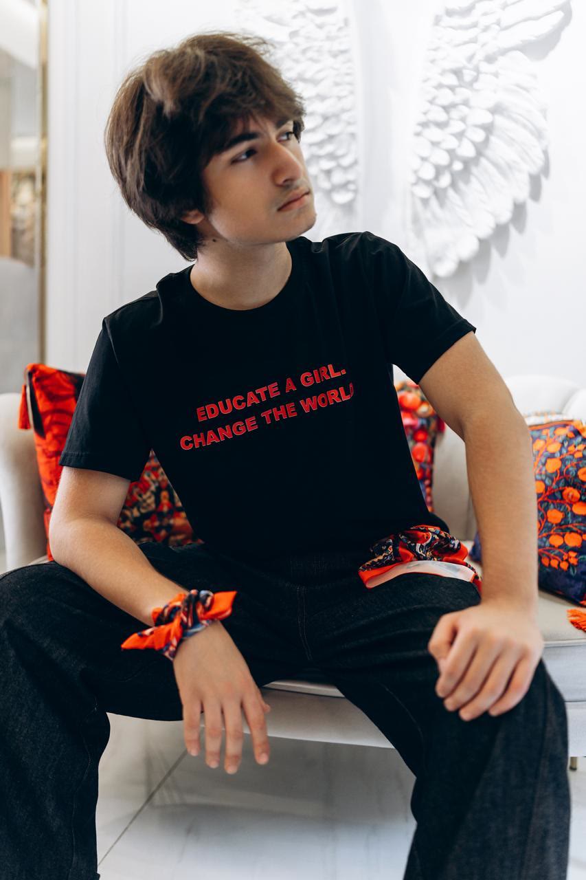Jamal sits on a white sofa among patterned cushions, in a black T-shirt that reads “Educate a girl. Change the world.”