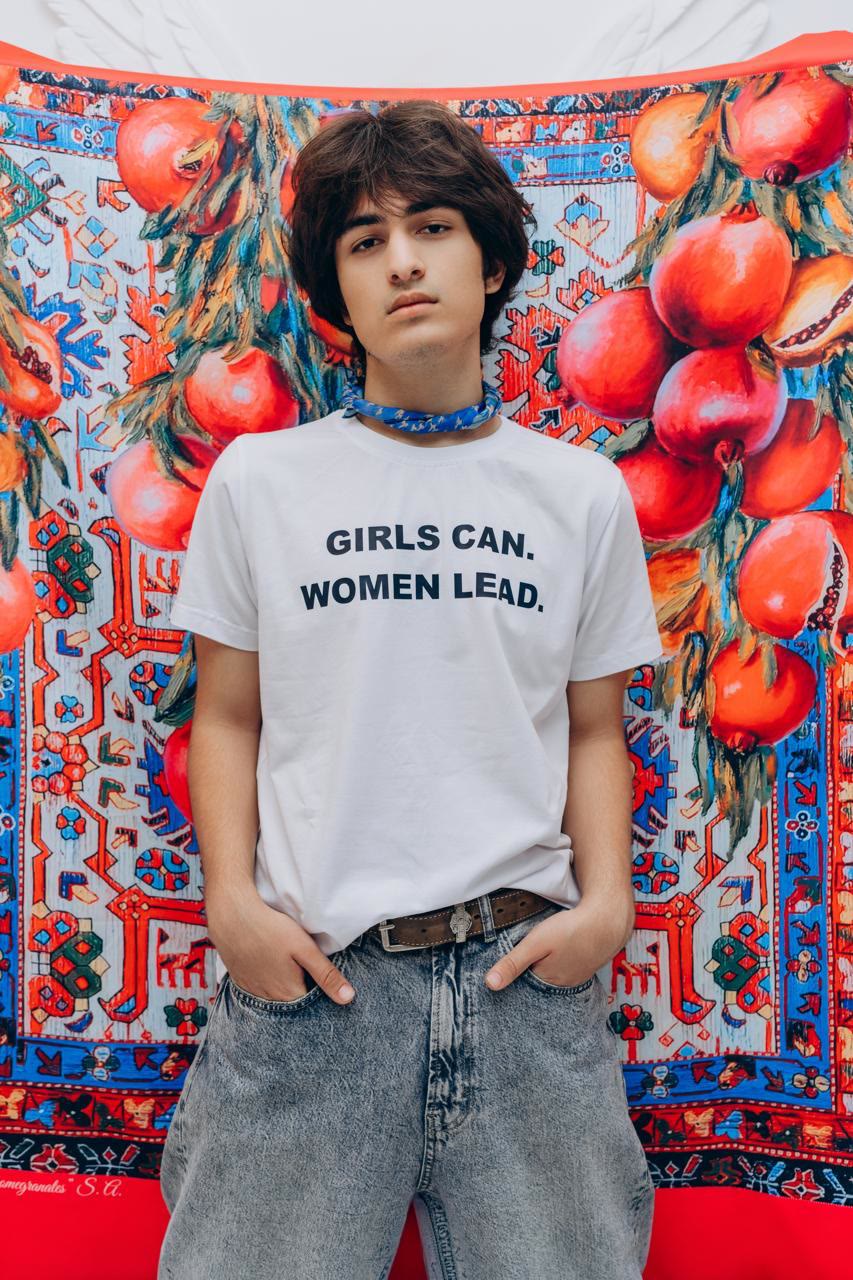 Jamal in a white T-shirt that reads “Girls can. Women lead.” in front of a painting of pomegranates over a carpet pattern.