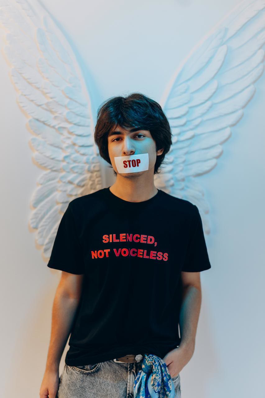 Jamal stands in front of white sculpted wings with a strip of tape marked STOP over his mouth, wearing a black T-shirt that reads “Silenced, not voiceless”.