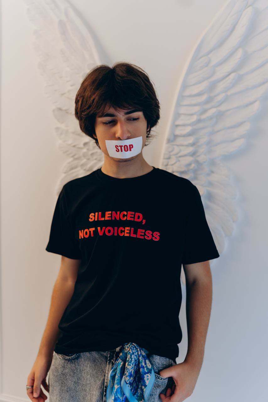 Jamal stands with his eyes closed in front of white sculpted wings, a strip of tape marked STOP over his mouth, in a black T-shirt that reads “Silenced, not voiceless”.