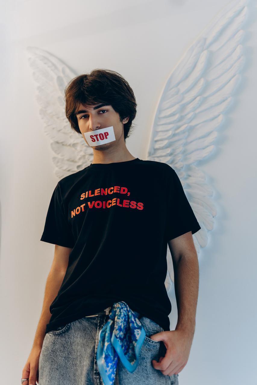 Jamal, seen from slightly below, with tape marked STOP over his mouth and white wings behind him.