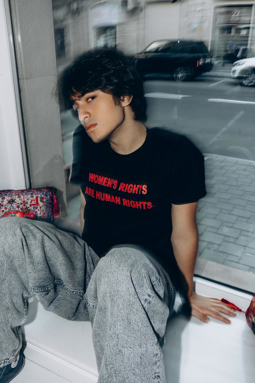 Jamal sits on a shop windowsill in a black slogan T-shirt, with the street blurred behind the glass.