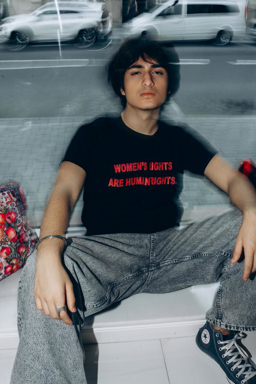 Jamal sits on a shop windowsill in a black T-shirt that reads “Women's rights are human rights” in red letters.